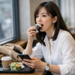窓辺のカウンターで、美人OLが短い昼休みにおにぎりを急いで頬張りつつスマホを確認している。コーヒーと小さなサラダがトレーに並ぶ。 A pretty office worker eats an onigiri quickly by a window counter while checking her phone, with coffee and a small salad on a tray.